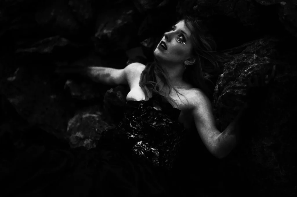 Jessie Monk avant‑folk artist reclining on rocky shoreline in black gown, eyes turned upward.